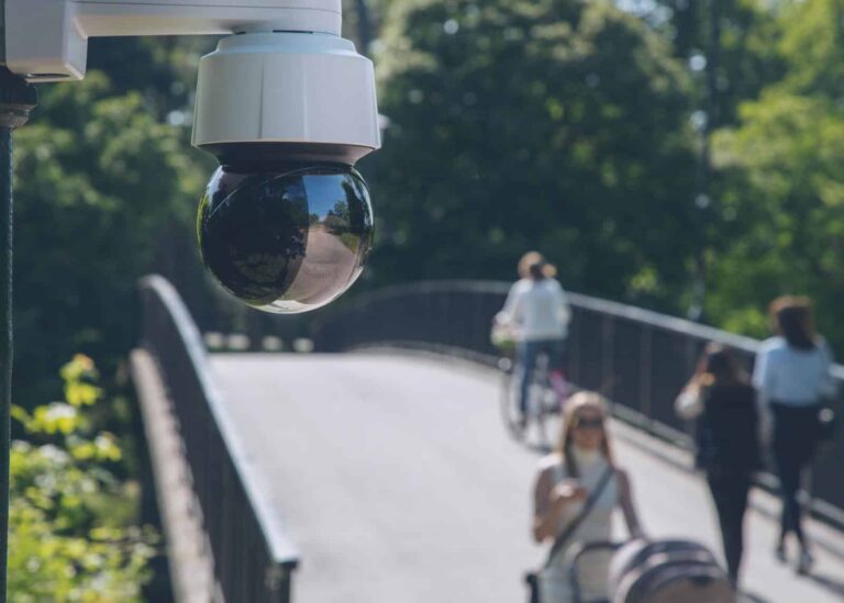 Axis Q6135-LE PTZ Surveillance camera overlooking a busy park bridge with pedestrians and cyclists on a sunny day, ensuring safety and monitoring activity.